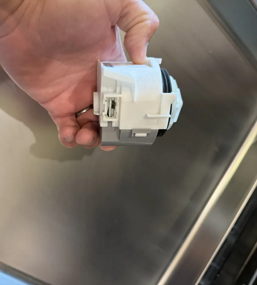 A dishwasher drain pump held in one hand above the stainless tub